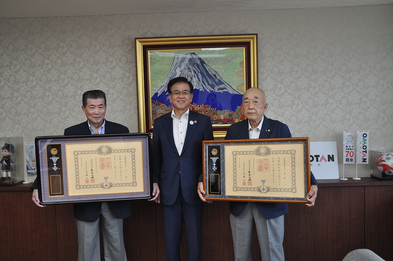 2人の男性が賞状のような大きな額縁を持ち、中央に立つスーツ姿の市長が笑顔でカメラに向かっている記念写真