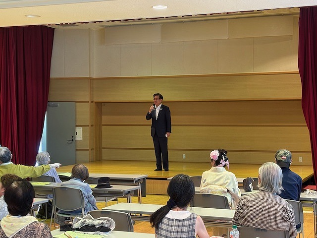 ステージ上でスーツを着た市長がマイクを持って話していて、参加者が椅子に座って聞いている写真