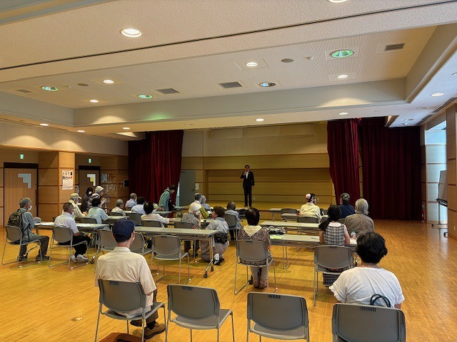 ステージ上にスーツの市長が立ちマイクを持ち話しており、前方の席には高齢者が複数座っている全体の様子を写した写真