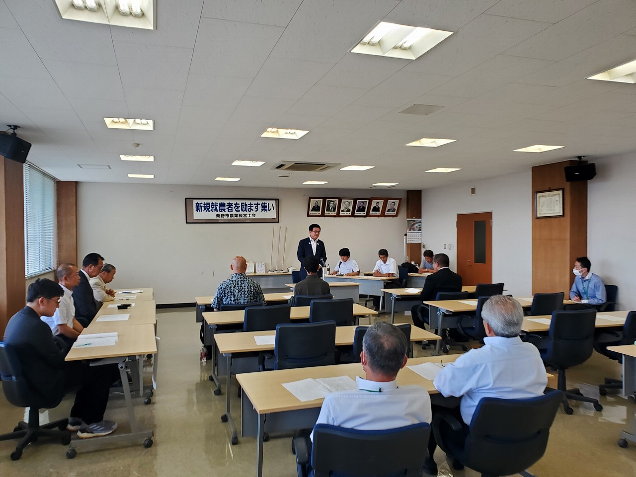会議室内で「新規就農者を励ます集い」と書かれた横断幕が掲げられた壁の前で、スーツ姿の市長が立って挨拶をしており、新規就農者や関係者が机に着いて真剣に話を聞いている様子の写真