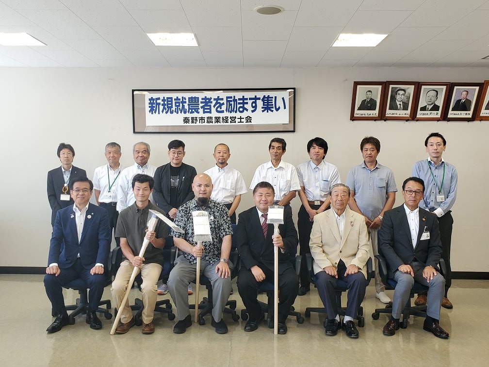 「新規就農者を励ます集い」と書かれた横断幕の前で、新規就農者と関係者が2列に並んで記念撮影をしており、前列中央の3人は鍬や看板など農業に関連する道具を手に持っている様子の写真