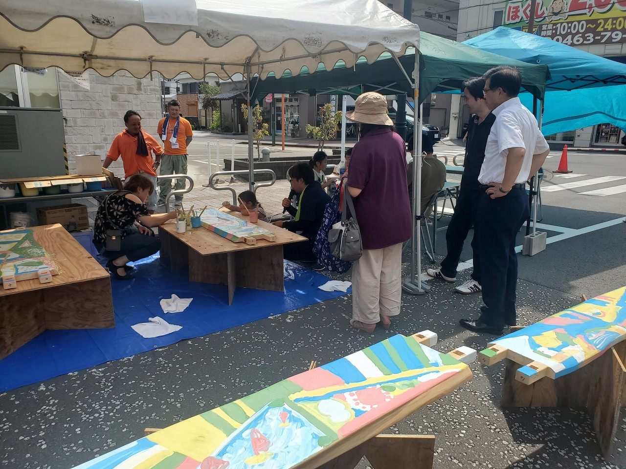 屋外のテントの下で木のベンチに色を塗るワークショップが開かれ、子どもたちが筆を使って熱心に絵を描いており、保護者や関係者が見守っている写真