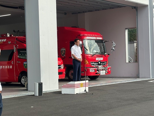 背後には「RESCUE」と書かれた赤い消防車が2台並んでおり、白いシャツと紺色のズボンを着た市長が小さな演壇に立ち挨拶をしている写真