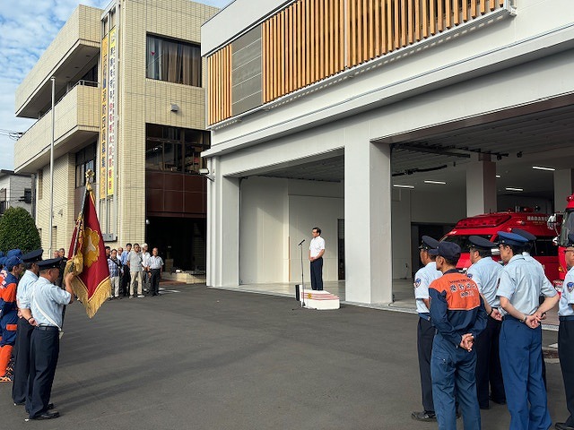 消防署の建物前で、白いシャツ姿の男性が演壇に立って挨拶をしており、手前には敬礼する制服姿の消防隊員たちや、旗を掲げて整列する隊員たちと関係者が並んでいる様子の写真