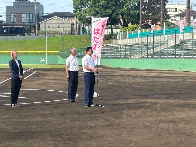 「女子野球タウンはだの」と書かれたピンク色の旗が立っており、スーツ姿の女性と男性がが整列し、市長がマイクを使って開会式のあいさつをしている写真
