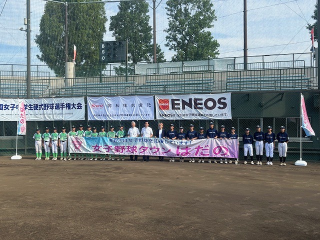 野球場のバックネット前にて、緑色と紺色のユニフォームを着た女子野球選手たちが横一列に並び、中央にいる関係者たちとともに「女子野球タウンはだの」と書かれたピンク色の横断幕を広げて記念撮影している写真
