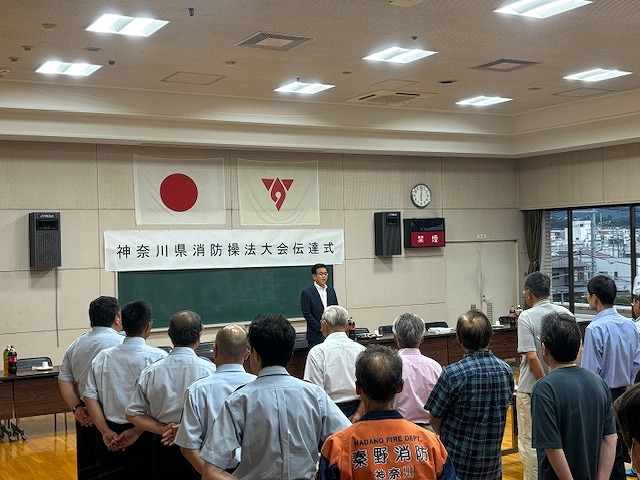 「神奈川県消防操法大会伝達式」の垂れ幕の前にスーツ姿の市長が立ち、整列して話を聞く消防関係者や市民に向けて挨拶をしている様子の写真