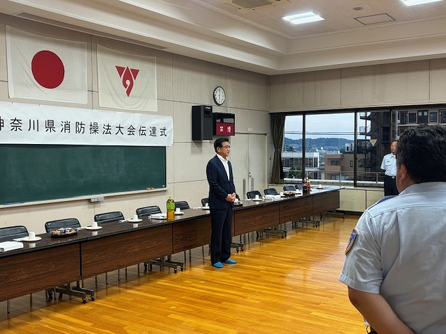 スーツ姿の市長が出席者に向けてまっすぐ立って話をしており、前には制服姿の消防職員が立って聞いている様子の写真