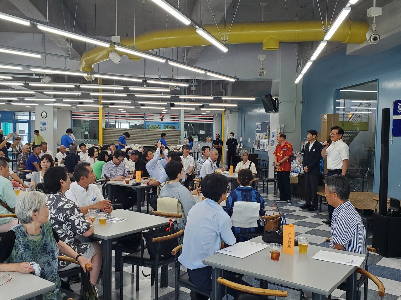 明るい照明の食堂で白いテーブルに着席した多数の参加者が、赤いシャツの講師や青いユニフォームのスタッフを囲んで説明を聞く地域交流イベントの写真