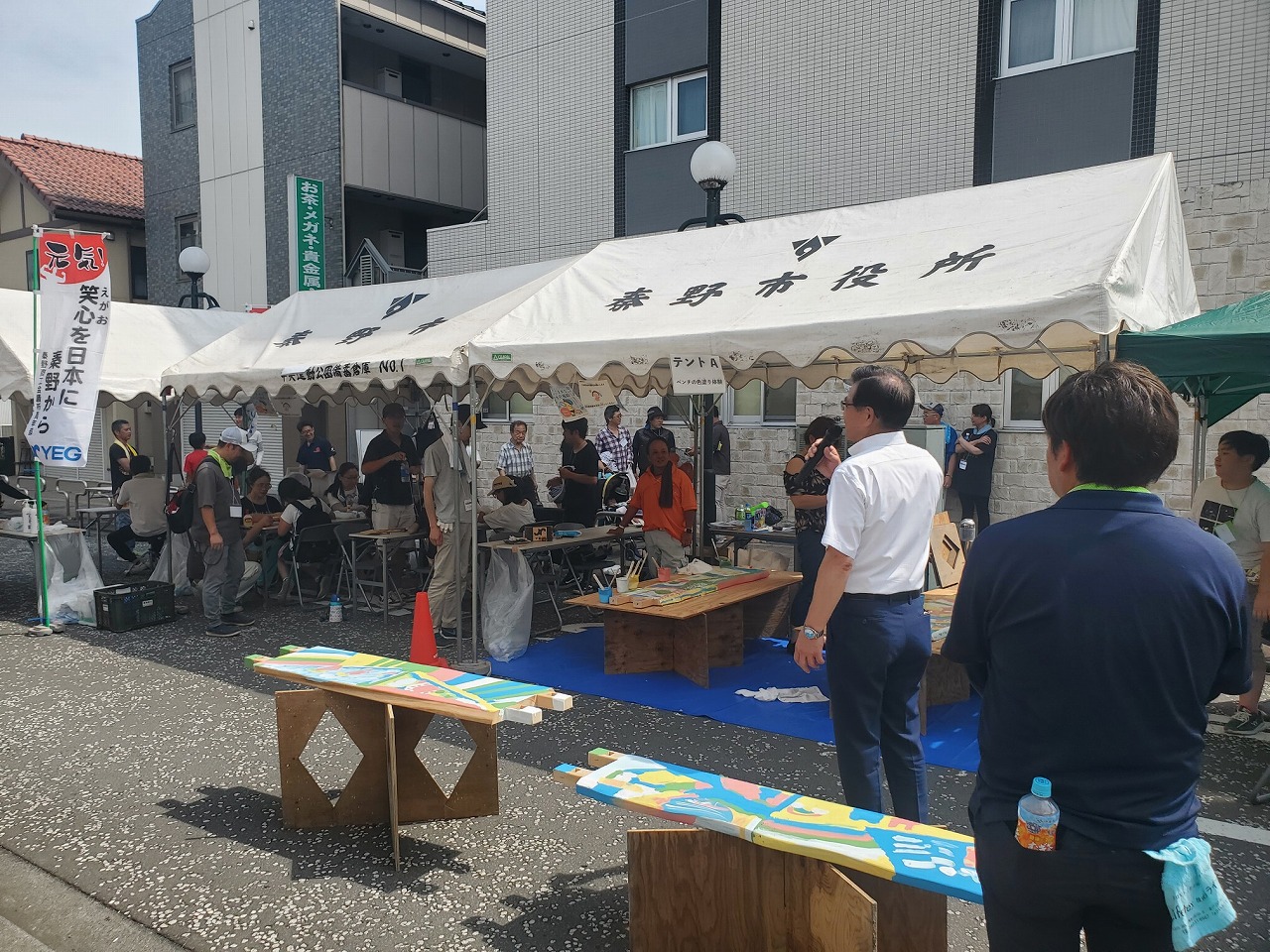 テントが設置された屋外の会場で、スーツ姿の市長がマイクを持って話をするのを多くの参加者が聞いている様子の写真