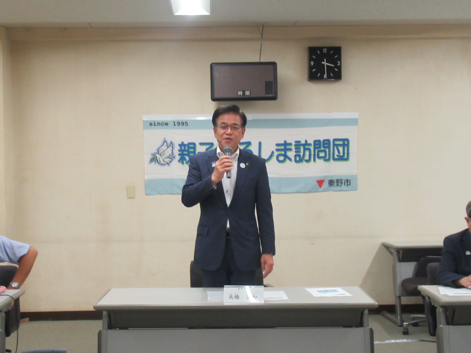 「親子のしま訪問団」と書かれた横断幕の前にマイクを手にスーツ姿で演台の前に立って話しをする市長の写真