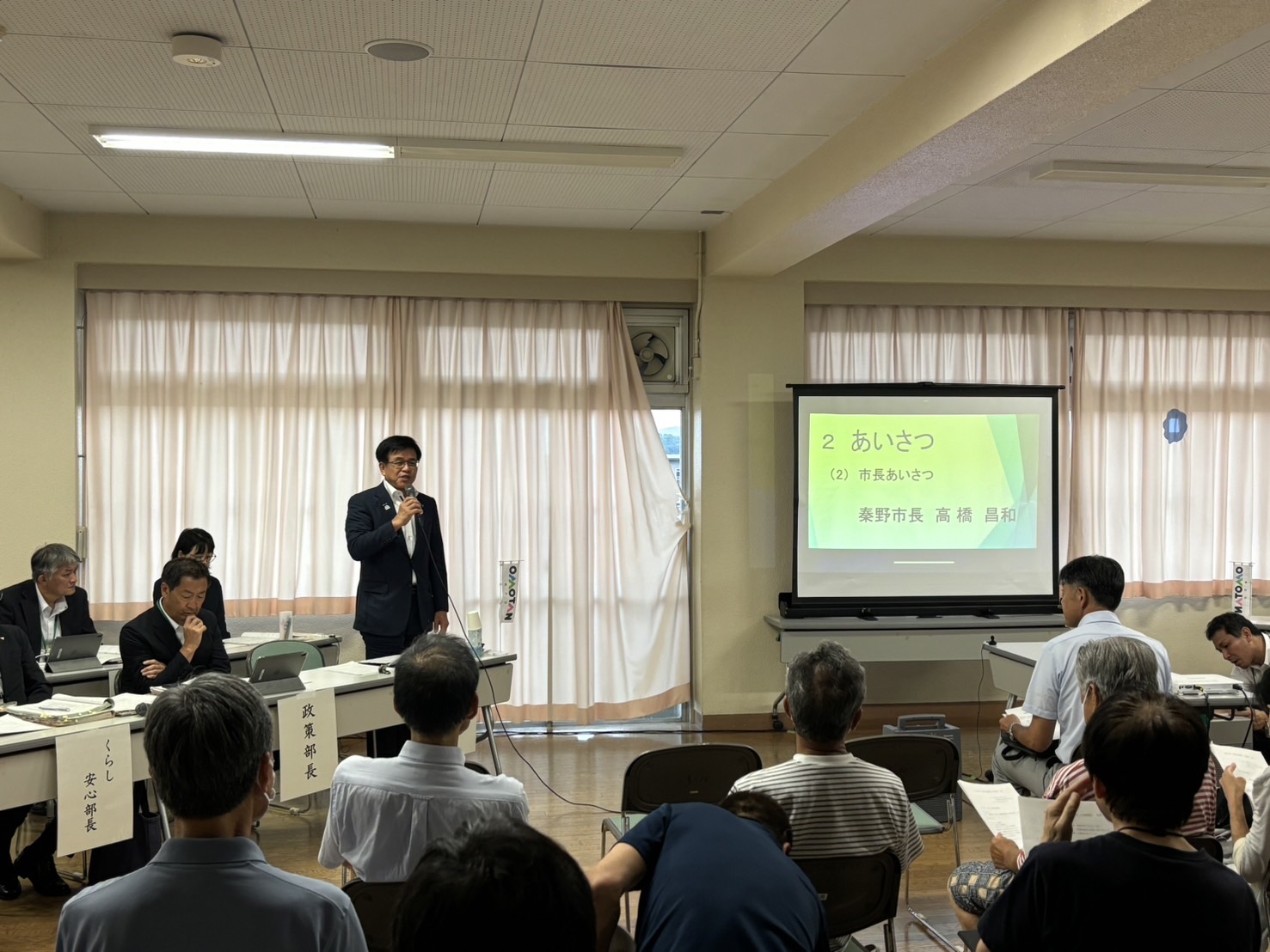 スクリーンに「市長あいさつ」と大きく表示され、市長が住民に向けて挨拶をしている地域懇談会の写真