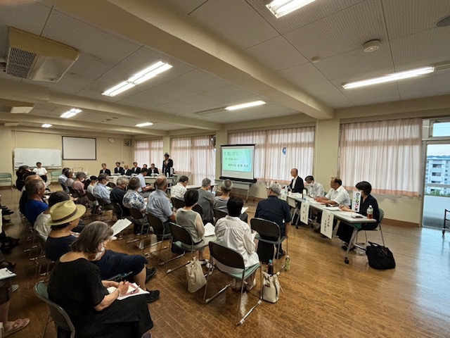 多数の住民が出席する中、市長がマイクを持ち、前方に並ぶ市職員とともにスクリーンを使用して地域課題について説明している、地域懇談会の全景の写真