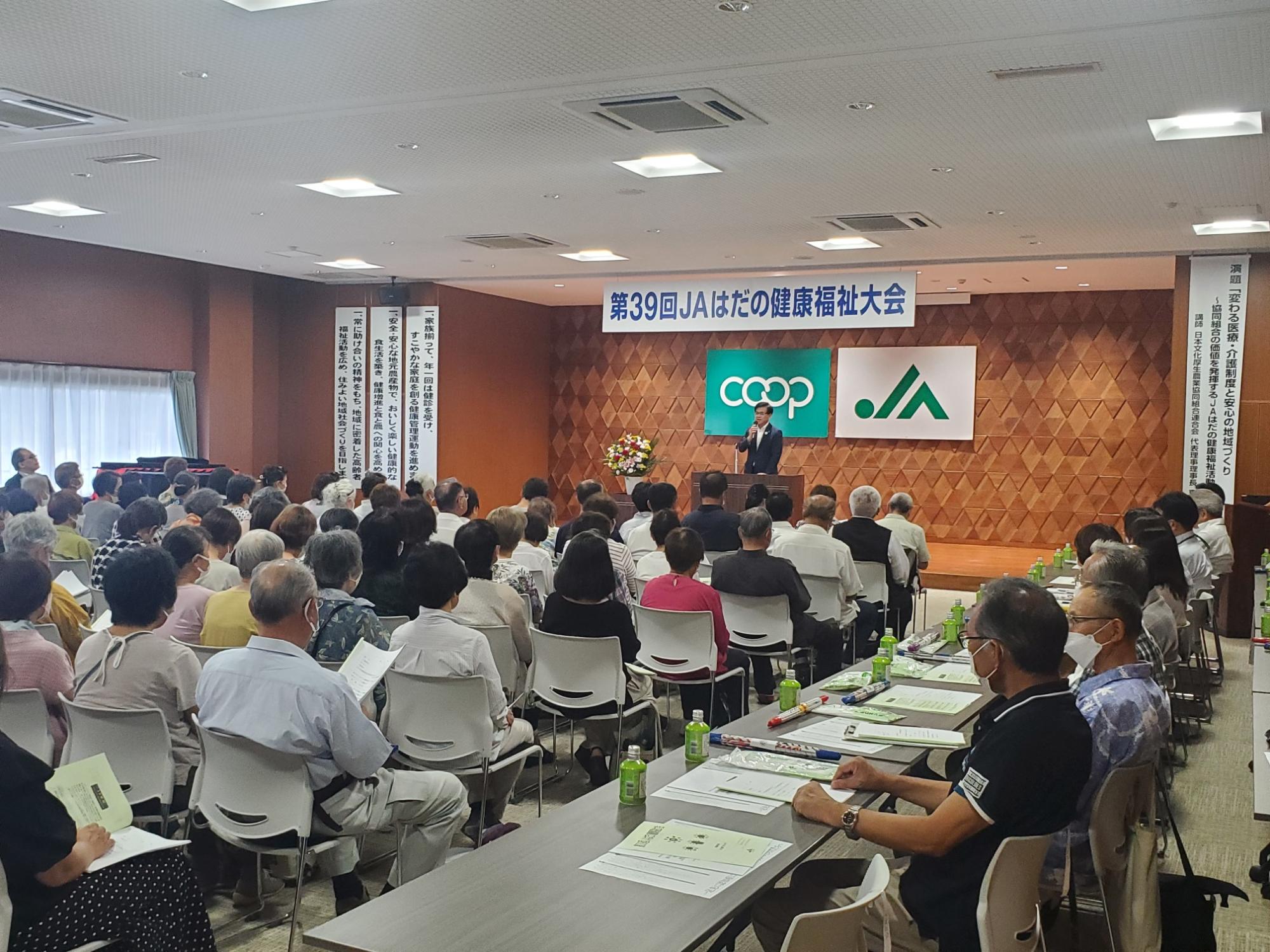 第39回JAはだの健康福祉大会の会場で、たくさんの参加者が椅子に座り、演台で話す市長の話を聞いている会場の様子の写真