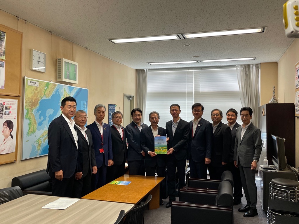 スーツ姿の複数の男性が中央の男性が持つ冊子に注目して並んでいる集合写真