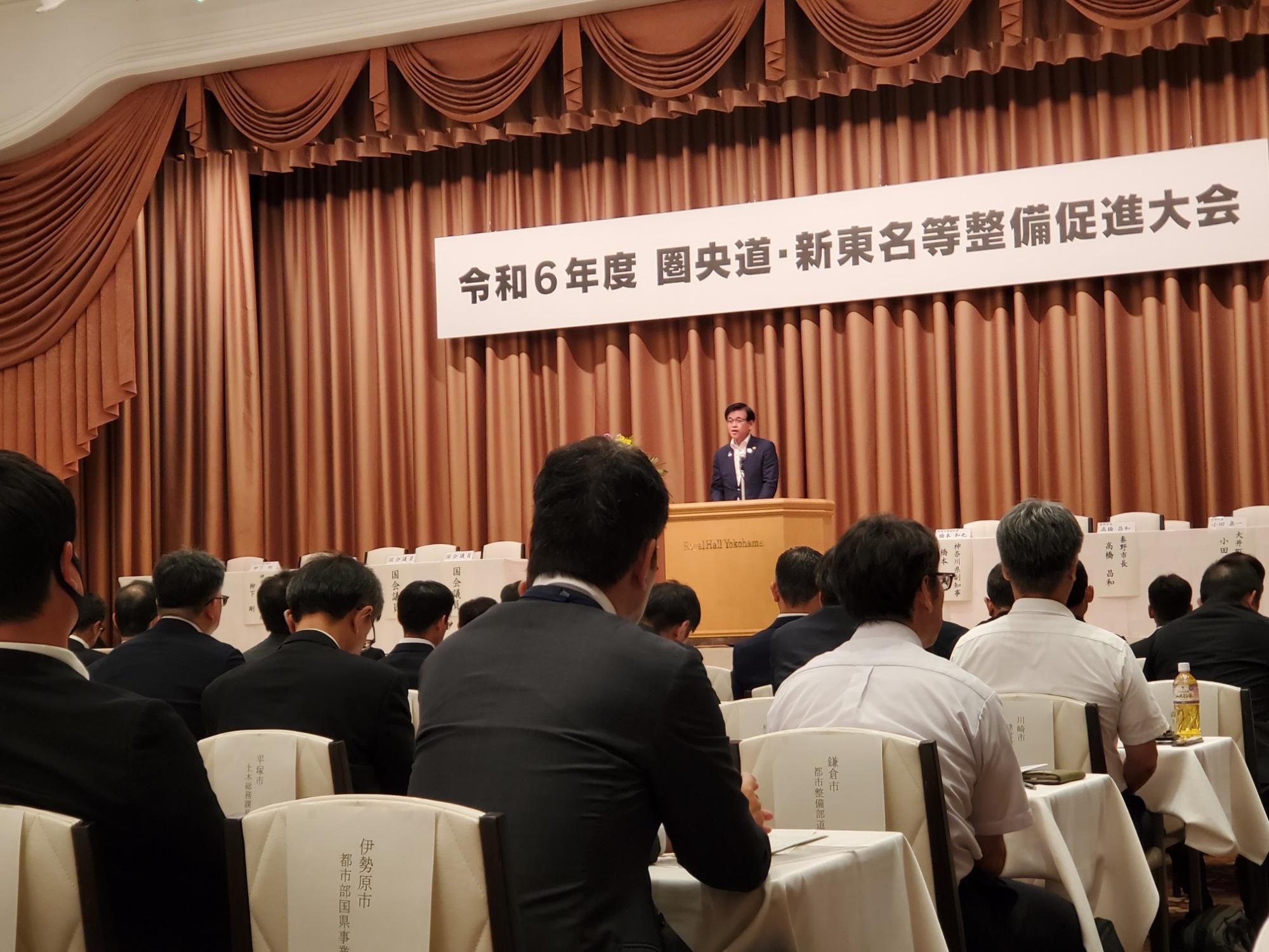 令和6年度 圏央道・新東名等整備促進大会の会場で、壇上の演台に立った市長が出席者に向けて演説を行っており、会場内の多くの関係者が着席して真剣に耳を傾けている様子の写真