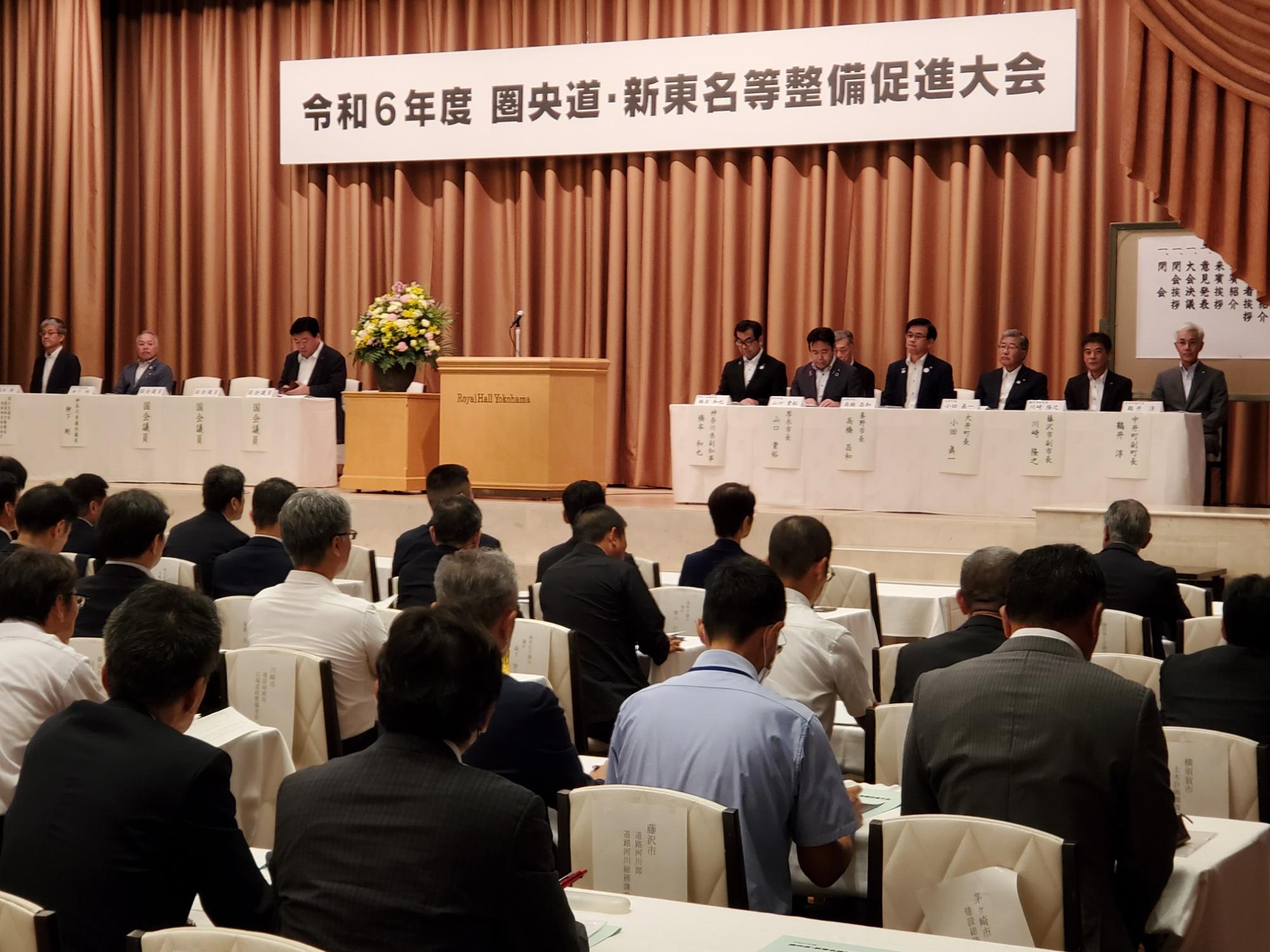 壇上に並んだ関係自治体や団体の代表者たちが席に座っており、正面には多数の来場者が資料を手にして話を聞いている様子の写真