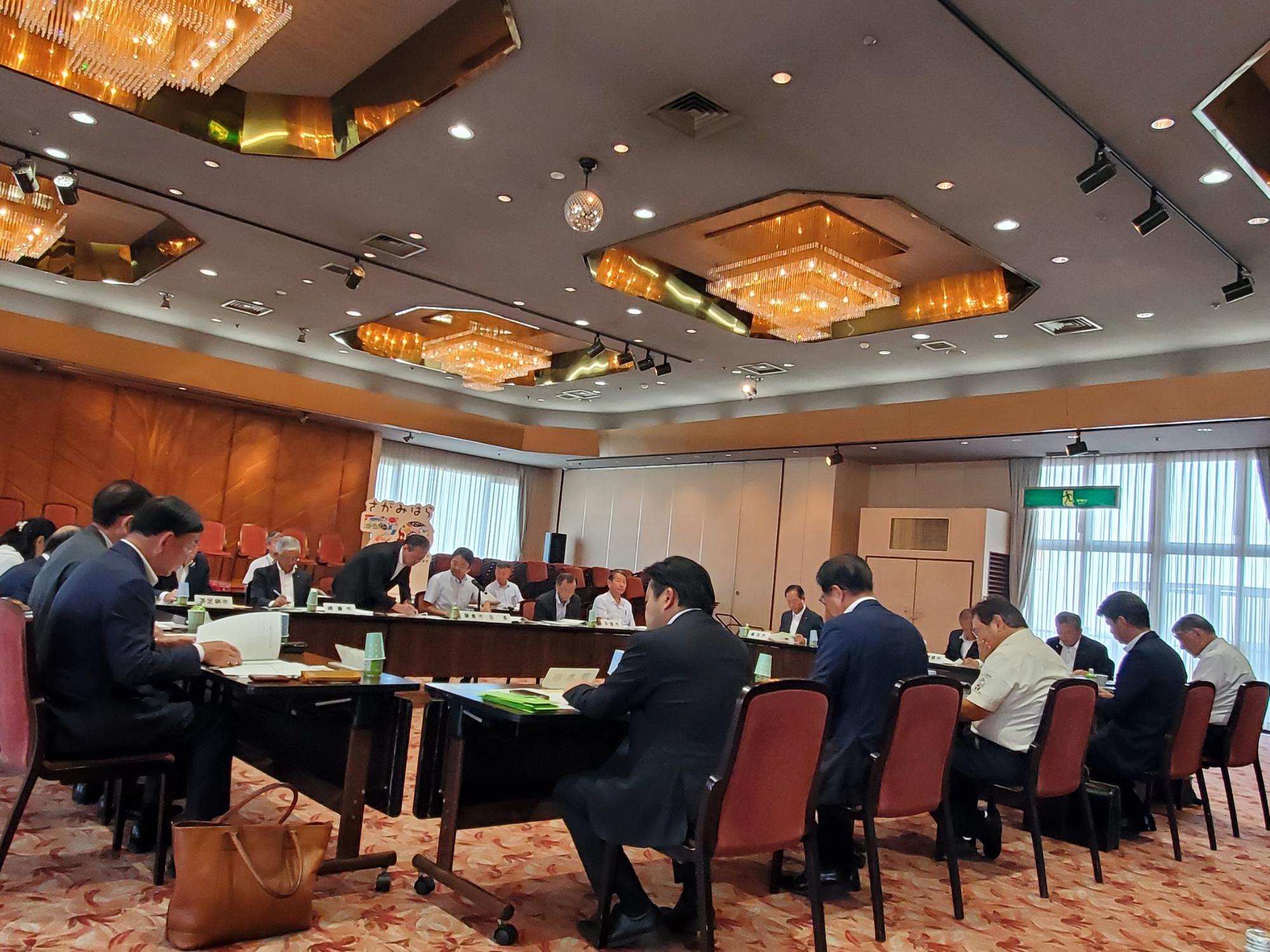 シャンデリアがある会議室で、多くのスーツ姿の参加者が長机を囲んで着席し、資料を確認しながら真剣な様子で会議を行っている様子の写真