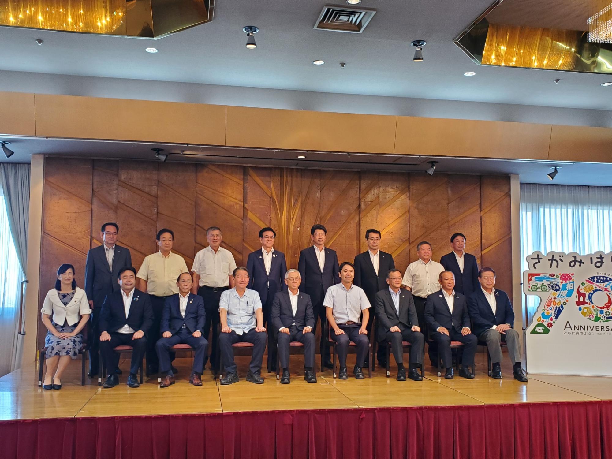 木目調の壁を背景に、スーツやビジネスカジュアル姿の男女が2列に整列し、「さがみはら 70周年」と書かれたカラフルなパネルの横で記念撮影をしている様子の写真