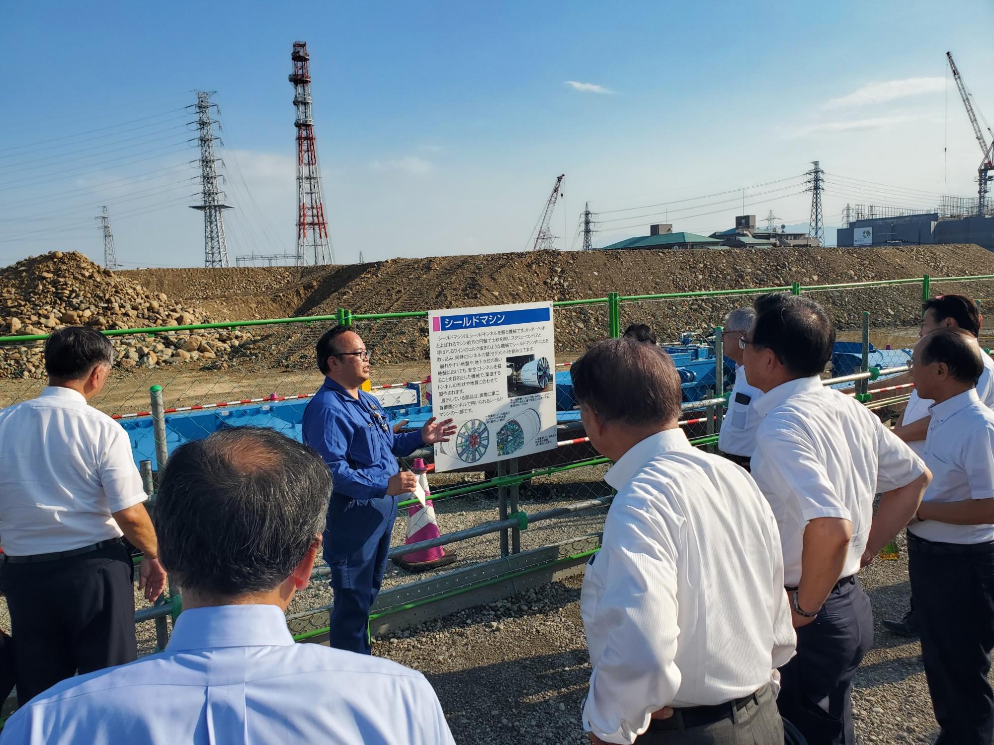 建設現場のような屋外で、青い作業着を着た男性が説明用パネルを使いながら、白いシャツ姿の視察団に向けて工事や施設について解説している様子の写真