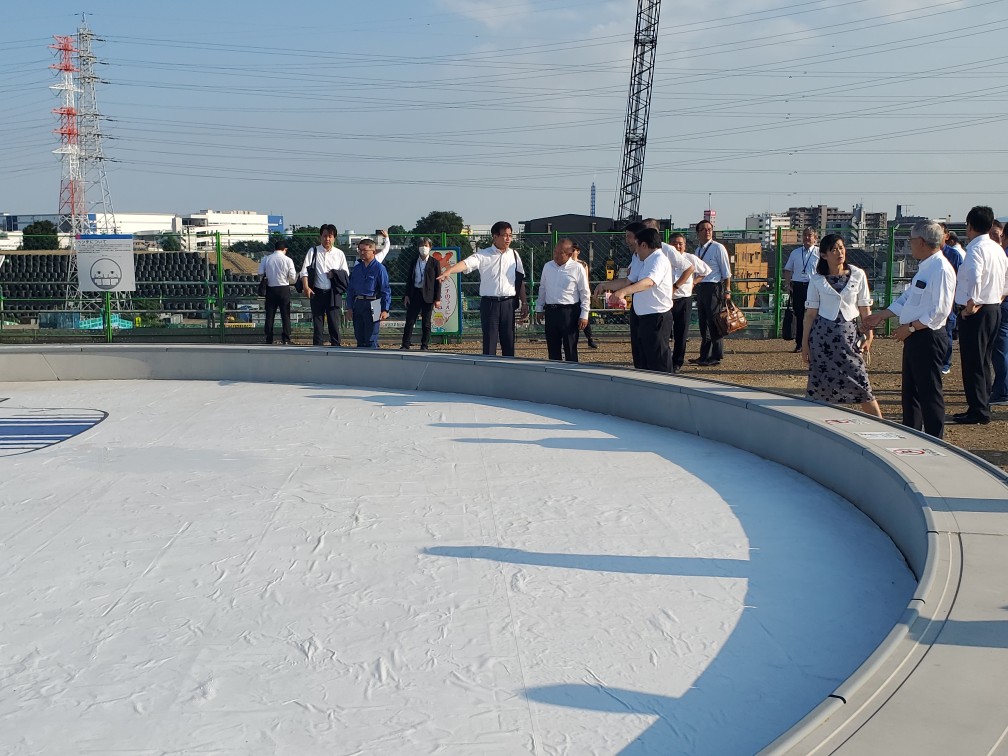 スーツや白シャツ姿の視察団が広い円形構造物の周囲に立ち、説明を聞いたり意見交換をしたりしている様子の写真