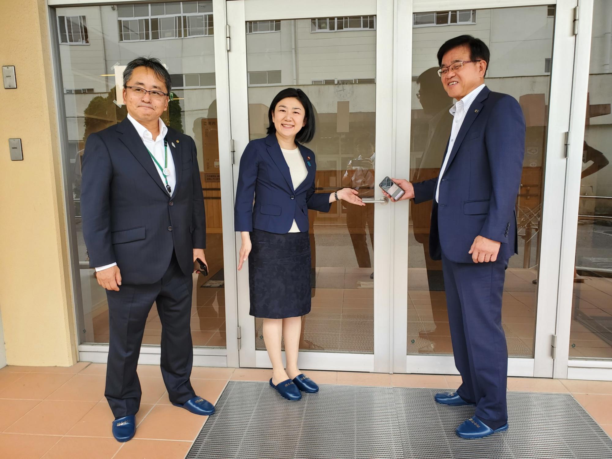 学校の正面玄関前で来訪者3人が笑顔でカメラに向かっている様子の写真