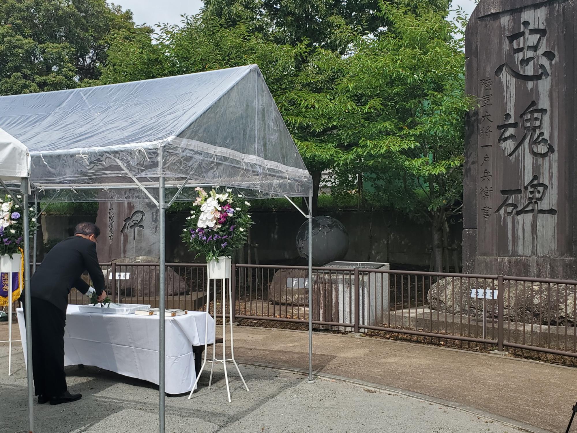 屋外に設置されている透明なテントの中に白い花が飾られ、スーツ姿の市長が献花台に花を添えている追悼式典の様子の写真