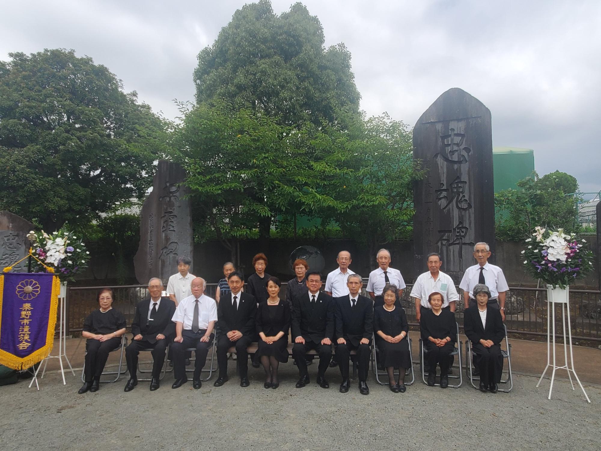 和装や喪服姿の男女が集まり、石碑を背景に、真剣な表情で座っている追悼式典の集合写真
