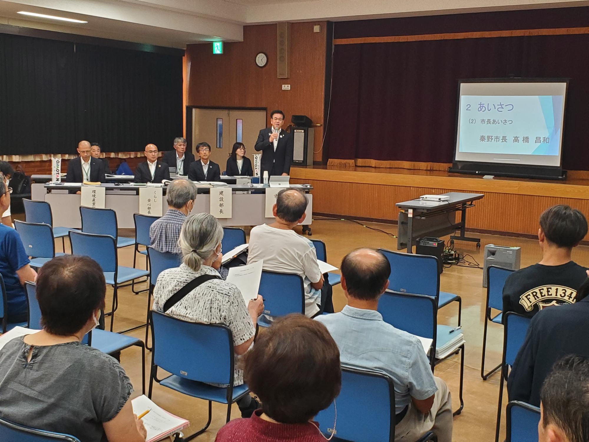 ステージのスクリーンに「あいさつ」と表示され、参加者が椅子に座ってスーツ姿の市長の挨拶を聞いており、前方には役員が並んでいる南地区の懇談会の様子の写真