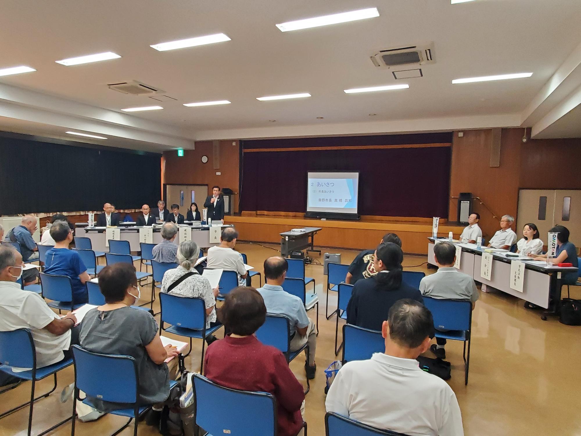 多数の参加者が椅子に座ってスーツ姿の市長の挨拶を聞いており、前方には複数の役員が並んでいる南地区の懇談会の様子の写真