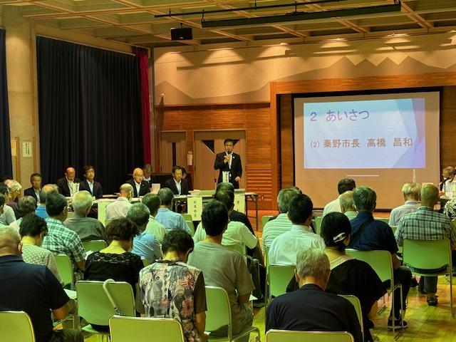 多数の参加者が椅子に座ってスーツ姿の市長の挨拶を聞いており、前方には複数の役員が並んでいる本町地区の懇談会の様子の写真