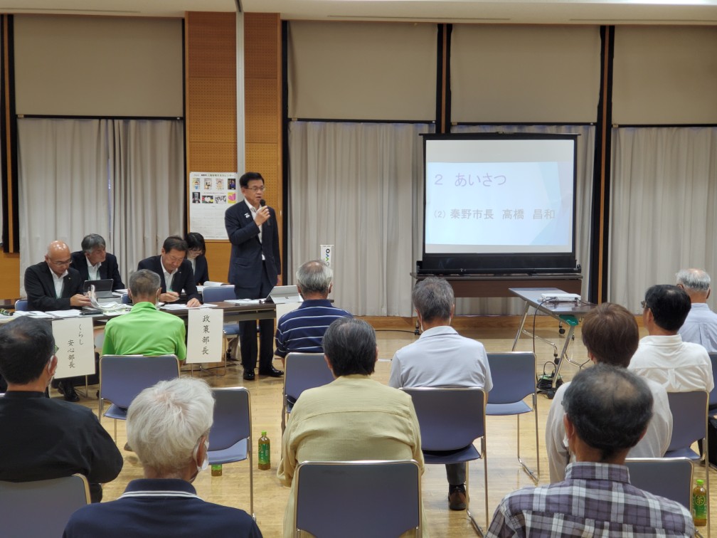 ステージ上でスクリーンに「あいさつ」と表示された前で、参加者が椅子に座り、スーツ姿の市長がマイクを持って聴衆に話しかけている様子の写真