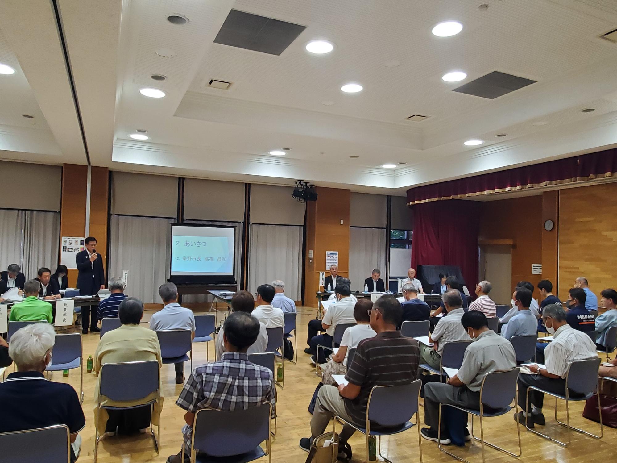 多数の参加者が椅子に座ってスーツ姿の市長の挨拶を聞いており、前方には複数の役員が並んでいる北地区の懇談会の様子の写真
