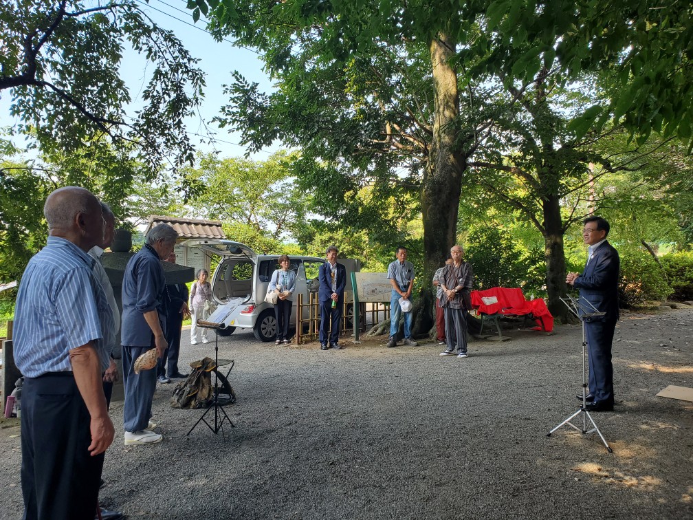 木陰の場所で、市長がマイクスタンドの前に立って挨拶を行い、住民や関係者が車の周辺や木の下に集まり話に耳を傾けている地域行事での挨拶の様子の写真