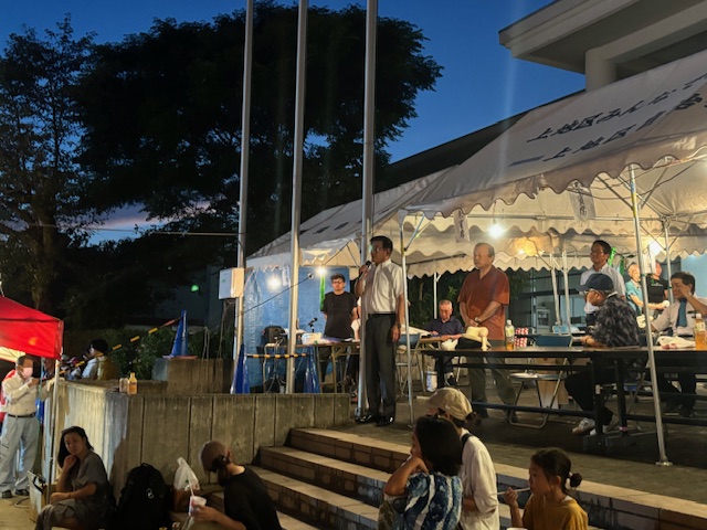 白いテントと提灯が並び、夜空の下で多数の人が集まっており、スーツ姿の市長が階段の上からマイクで挨拶をしている様子の写真