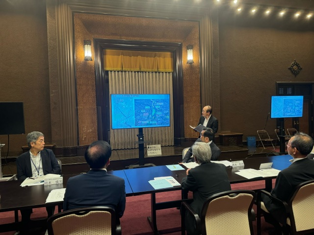 会議室で、前方のスクリーンに資料を参加者に見せながら、スーツ姿の男性がプレゼンテーションを行っている会議の様子の写真