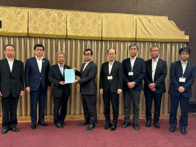 光沢のあるカーテンを背景に、スーツ姿の男性が横一列に並んでおり、中央の2人が青い表紙の冊子を両手で持ち、カメラに向かっている記念写真