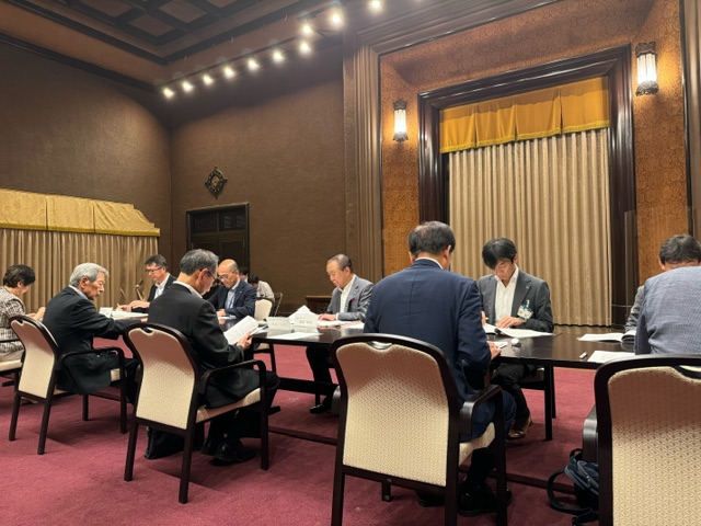 落ち着いた照明の会議室で、テーブルを囲んで多数の参加者が座っており、皆が資料に目を通している会議の様子の写真