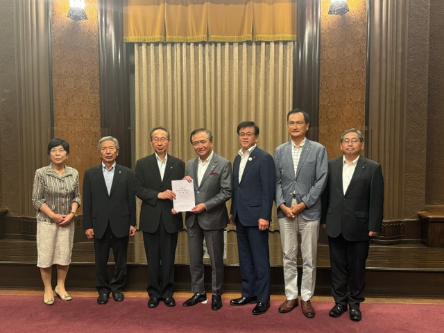 スーツ姿とビジネスカジュアルの服装の関係者が並び、中央の2人が書類を手に持ち記念撮影をしている写真