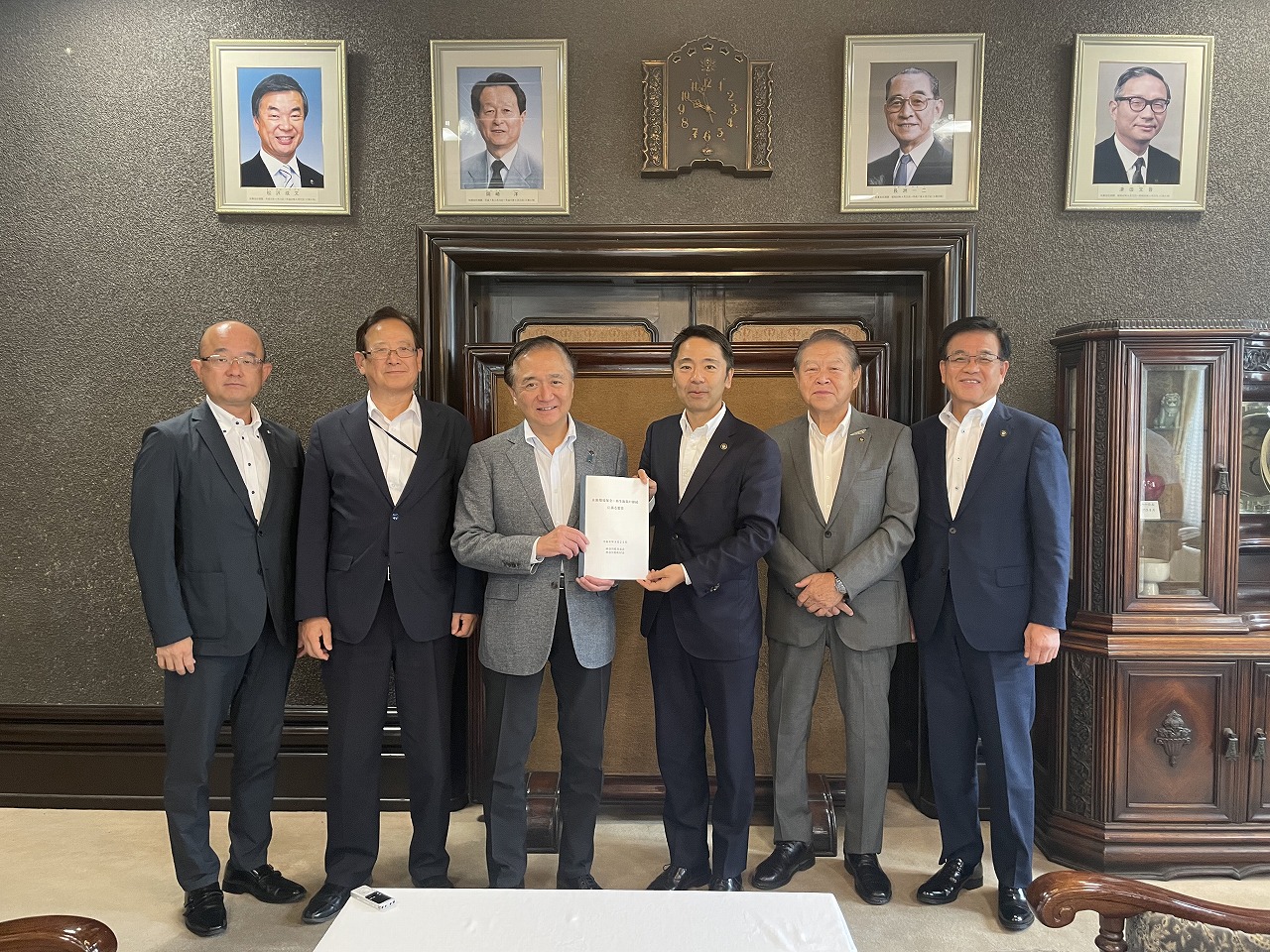 スーツ姿の6人の男性が並び、中央の2人が書類を持ってカメラに向かっている記念写真