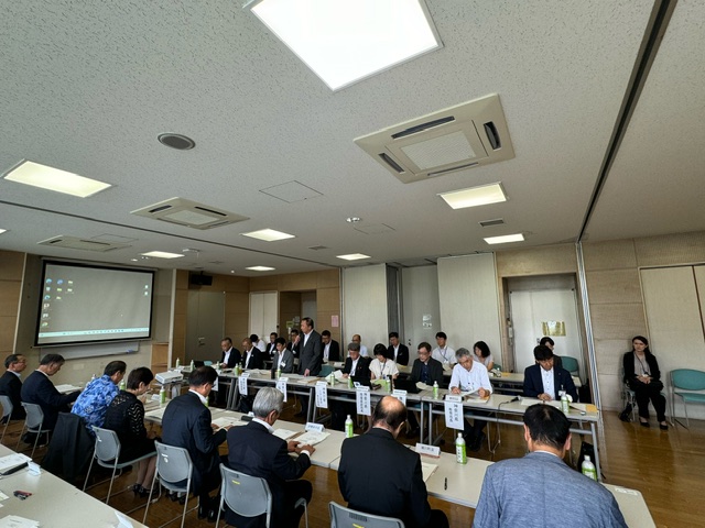 会議室で複数の参加者が机に座り、中央に立つスーツ姿の男性が、資料を確認しながら話している様子の写真