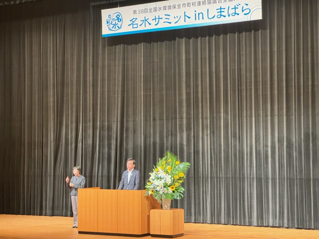 「名水サミット in しまばら」の横断幕が掲げられた舞台で、演台に立つスーツ姿の市長が挨拶をしており、隣に手話通訳者が立っている写真