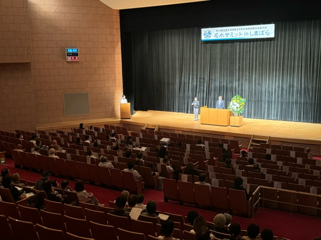 「名水サミット in しまばら」の横断幕が掲げられた舞台で、演台に立つ市長と手話通訳者が登壇しており、客席には多くの来場者が資料を手にして着席している会場全体の様子の写真