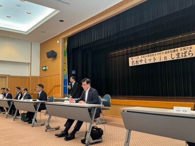 「名水サミット in しまばら」の横断幕が掲げられた会場で、複数のスーツ姿の出席者が机に着席し、資料を確認しながら協議や発言を行っている、会議中の様子の写真