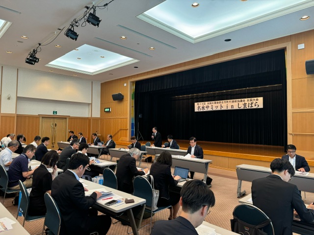 「名水サミット in しまばら」の横断幕が掲げられたホールで、複数の自治体関係者が長机に着席し、資料を読みながら真剣な表情で会議に参加している会議風景の写真
