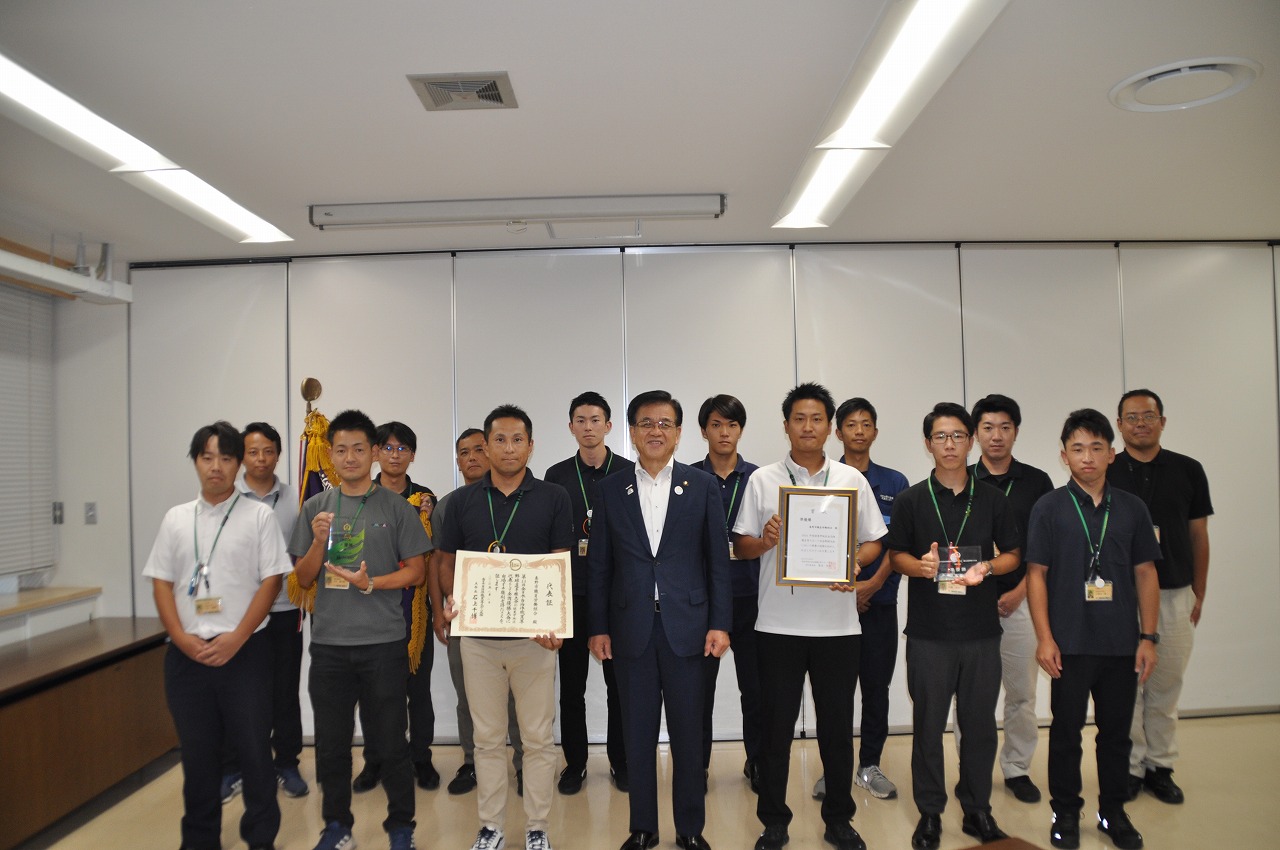 表彰状やトロフィーを手にした複数の男性職員とスーツ姿の市長を中心に整列している笑顔の集合写真