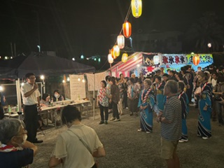 夜の屋外会場で、提灯の灯りの下、多くの来場者が集まる中、浴衣姿の踊り手や関係者が並び、市長がマイクで挨拶をしている様子の写真