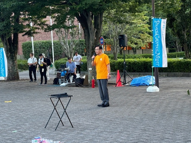 屋外の広場で、オレンジ色のTシャツを着た市長がマイクを手にスピーチをしている写真