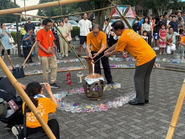 円形に並べられたグラスの中心に置かれたキャンドルを灯す男性たちの様子を、多くの観客が囲んで見守っている、キャンドルナイトイベントの写真