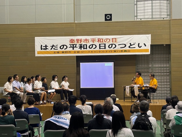 「秦野市平和の日はだの平和の日のつどい」と書かれた横断幕の下、大人と子どもたちが着席した向かい側に黄色のTシャツを着た二人がし座り話をしている写真